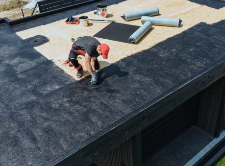 Roofer installing EPDM roofing membrane on a flat commercial roof using specialized tools and adhesive.